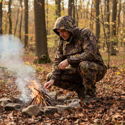Person in camouflage attire tending to a campfire in a forest setting