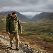 Man hiking on a mountain trail with a backpack, surrounded by rugged mountains, wearing jackpyke galbraith smock